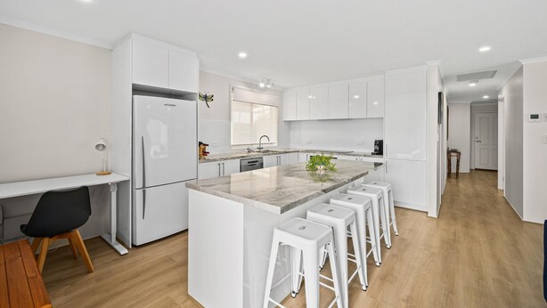 A modern kitchen with white cabinetry, a central island with bar stools, a refrigerator, and a dining area with a table and chairs. - Aldinga Rose - Aldinga Beach - C21 SouthCoast Holidays (Aldinga Beach)