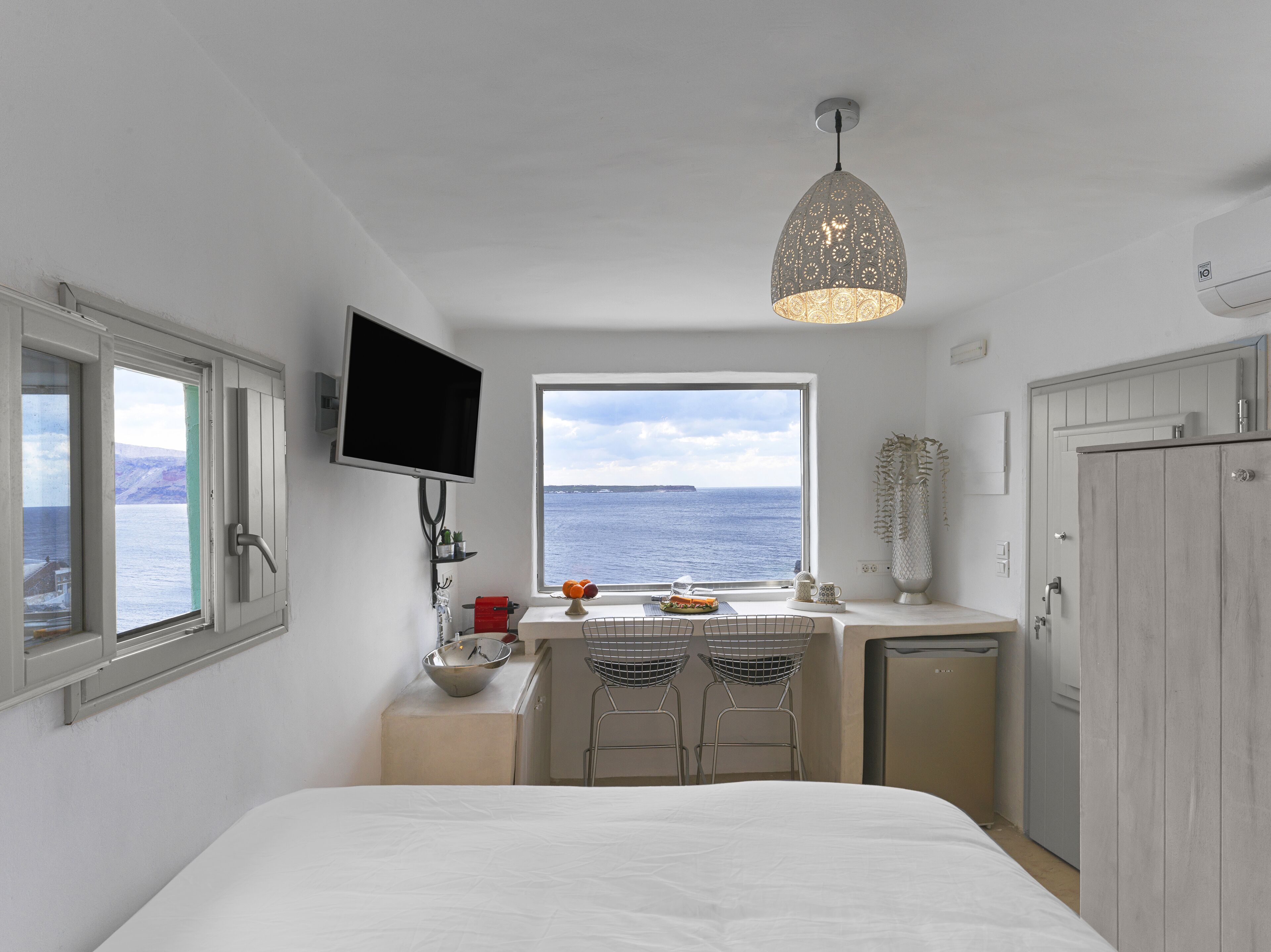 Ein Schlafzimmer mit Bett, Wand-Fernseher, Fenster mit Meerblick und einer Küchenzeile mit Spüle und Stühlen.