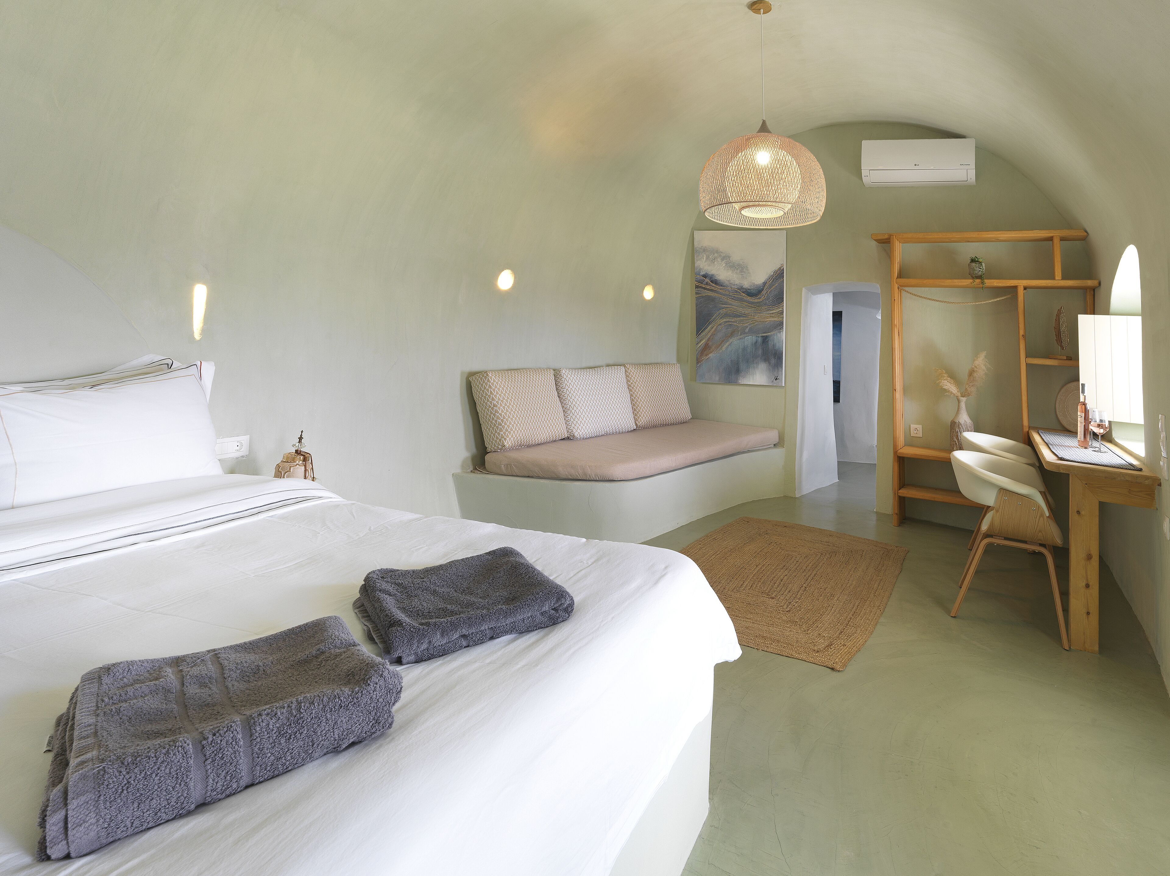 A bedroom with a large bed, a bench, a desk, and a hanging light fixture.