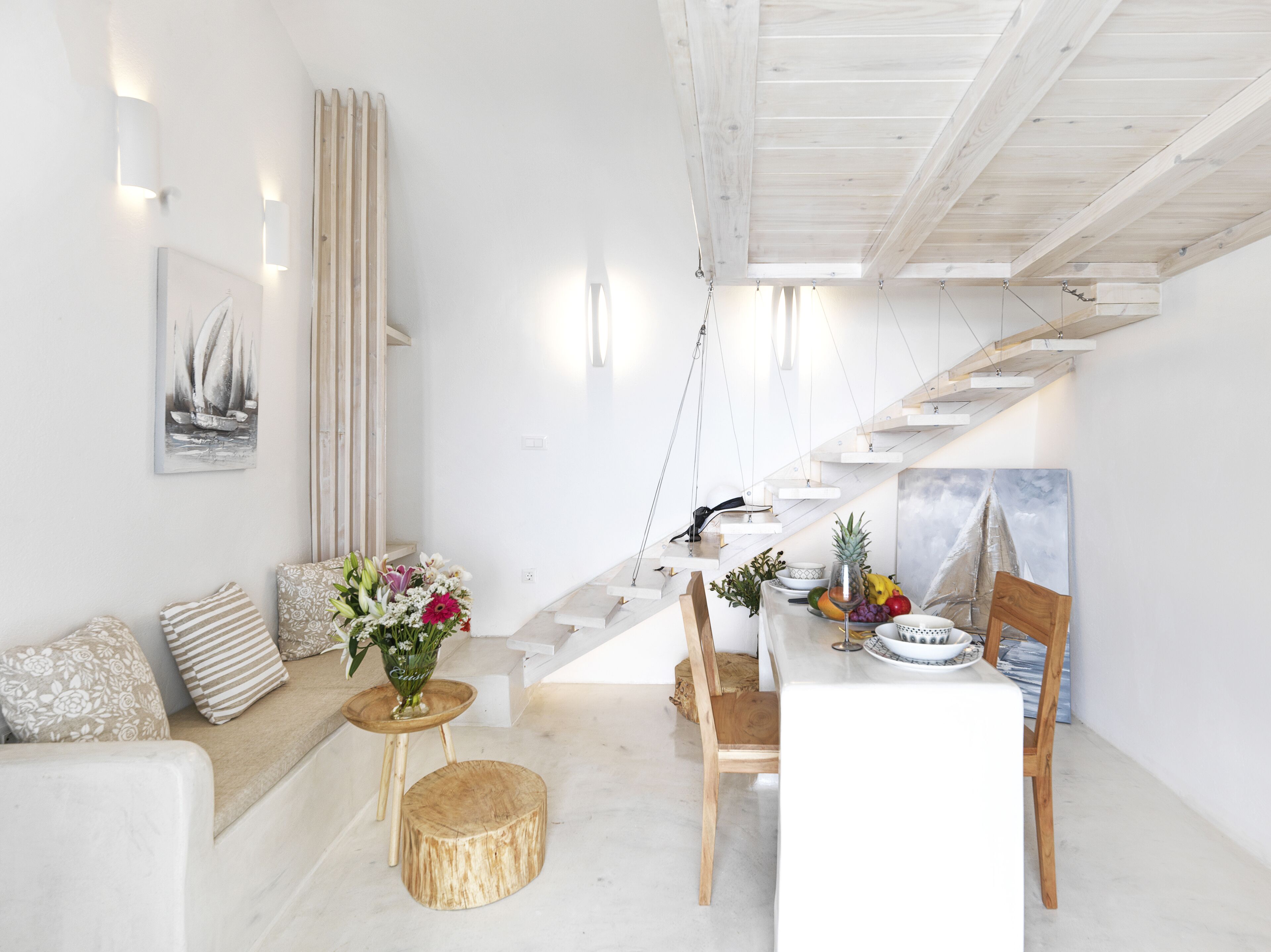 Um interior moderno e bem iluminado, com uma escada que leva a um andar superior, uma área de jantar com uma mesa posta para dois e um espaço de estar com um sofá e almofadas.