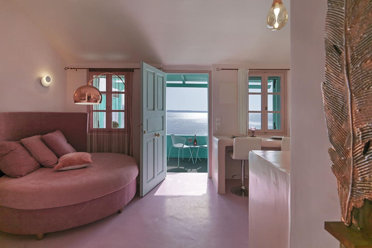 A bedroom with a pink sofa, a desk with a chair, a window with a view of the sea, and a pendant light.