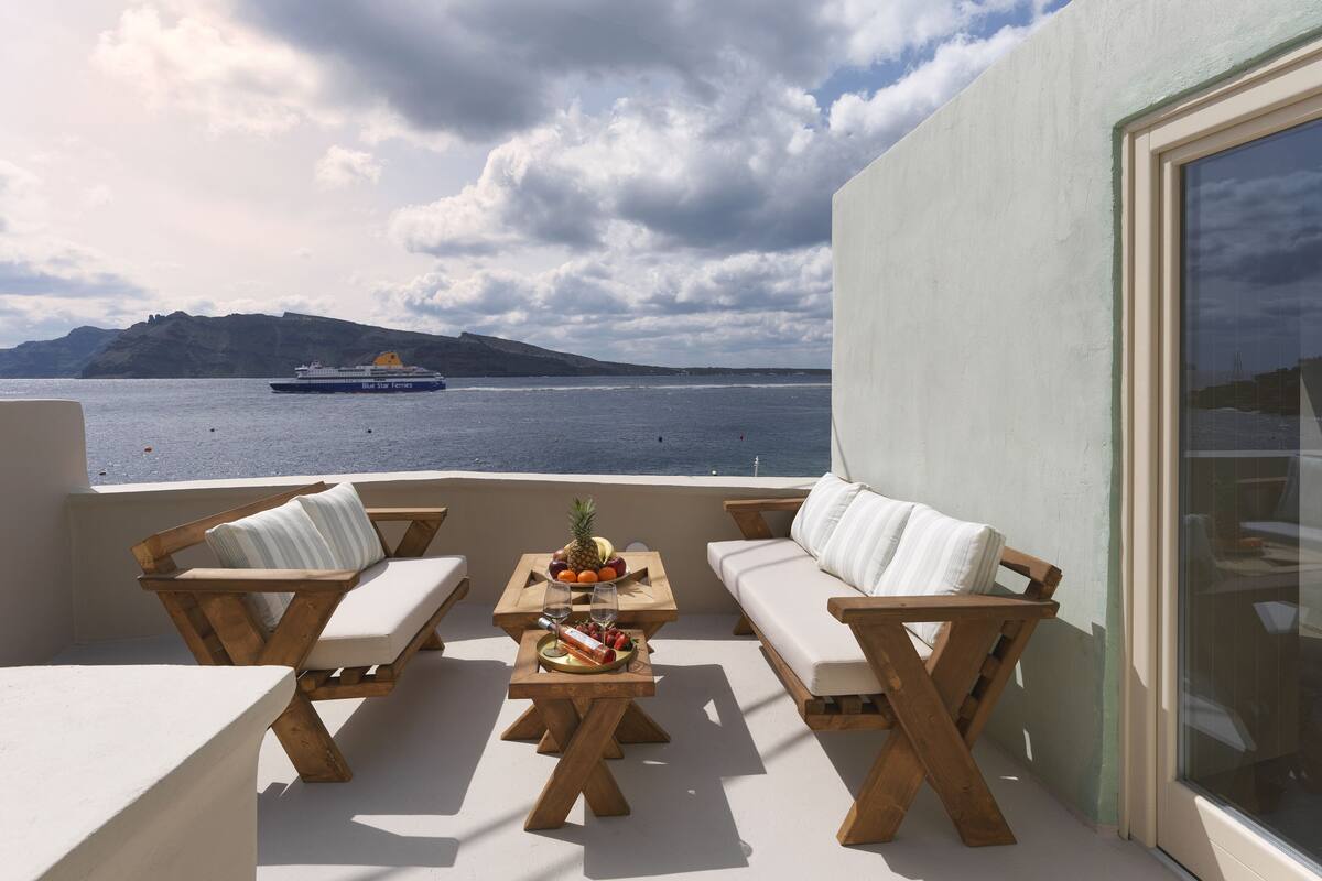 A balcony with wooden furniture, a table with fruits and glasses, and a view of the sea with a ferry.