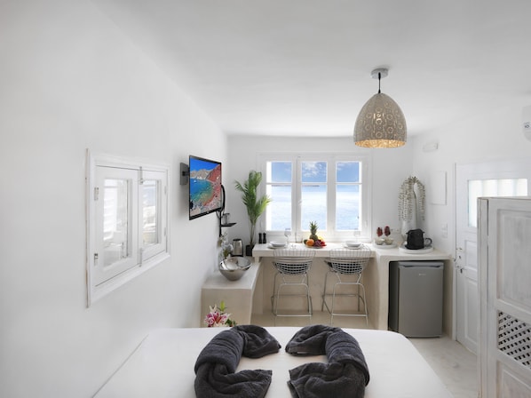 A compact hotel room with a bed, a small kitchenette, a dining area with two chairs, a TV mounted on the wall, and a large window offering a view of the sea. - Secret Gem Suites (Santorini)