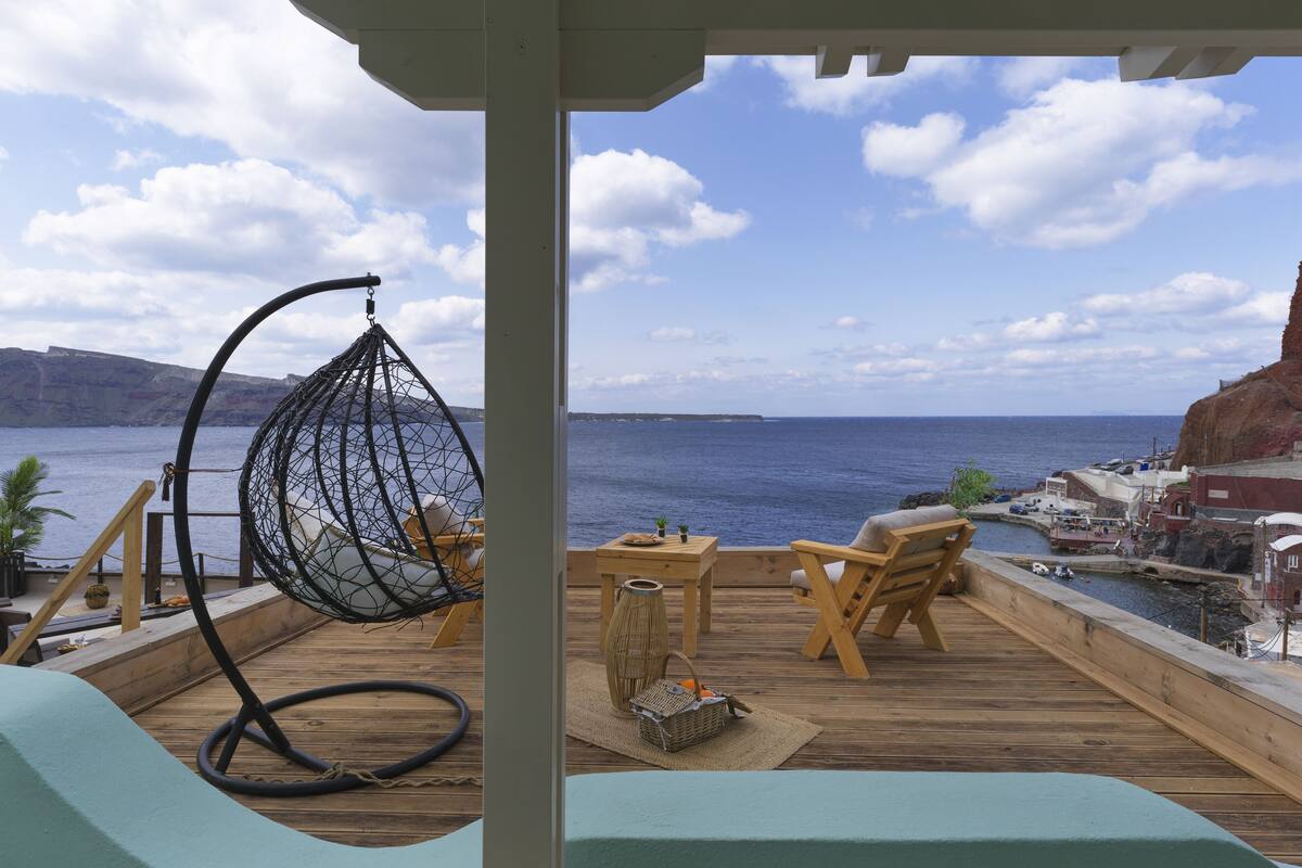 A wooden deck with a hanging chair, a table, and a view of the sea and cliffs.