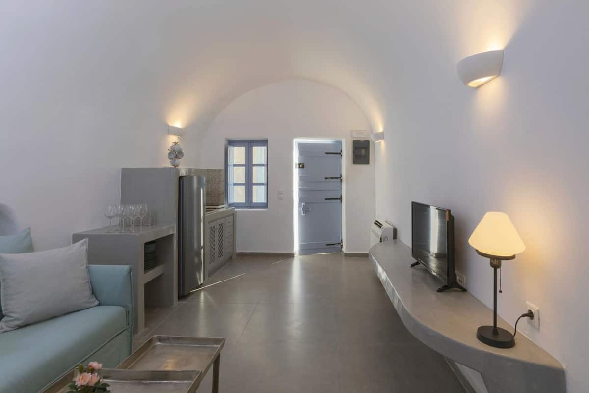 A modern living room with a sofa, a coffee table, a TV, and a lamp. There is a kitchen area with cabinets and a refrigerator.