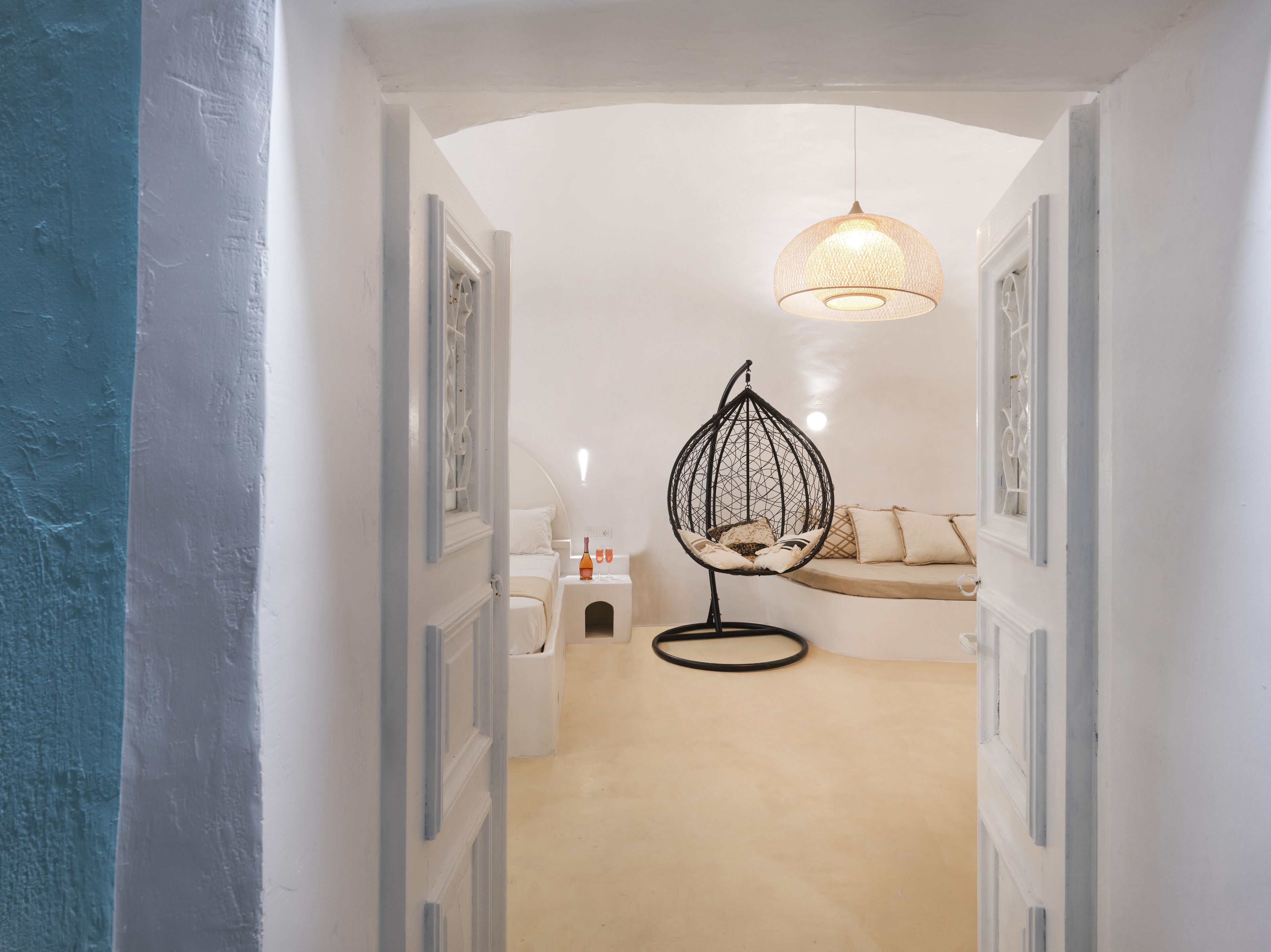 Een gang die leidt naar een slaapkamer, met een hanglamp, een hangstoel en een nachtkastje met een fles en glazen.