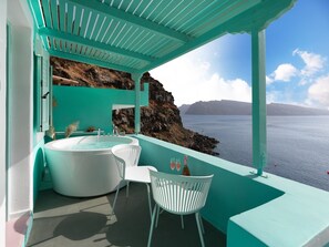 A modern bathroom with a large bathtub, white chairs, and a view of the sea. - Secret Gem Suites (Santorini)