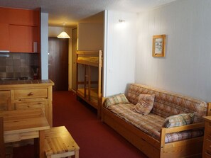 A compact room with a kitchenette, a wooden bench, a sofa with plaid bedding, and a framed picture on the wall.