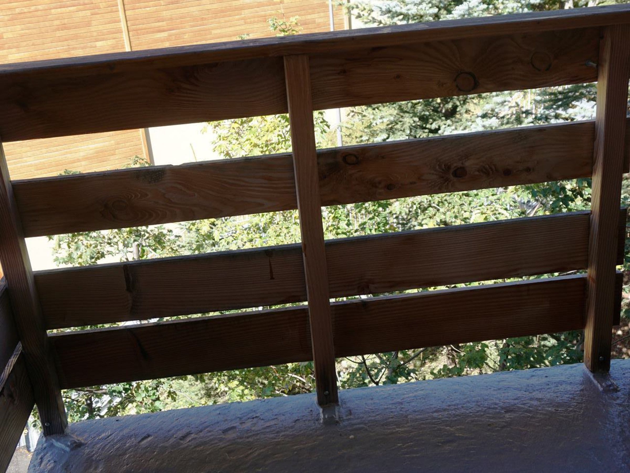 A wooden balcony railing with a view of trees and a building in the background.
