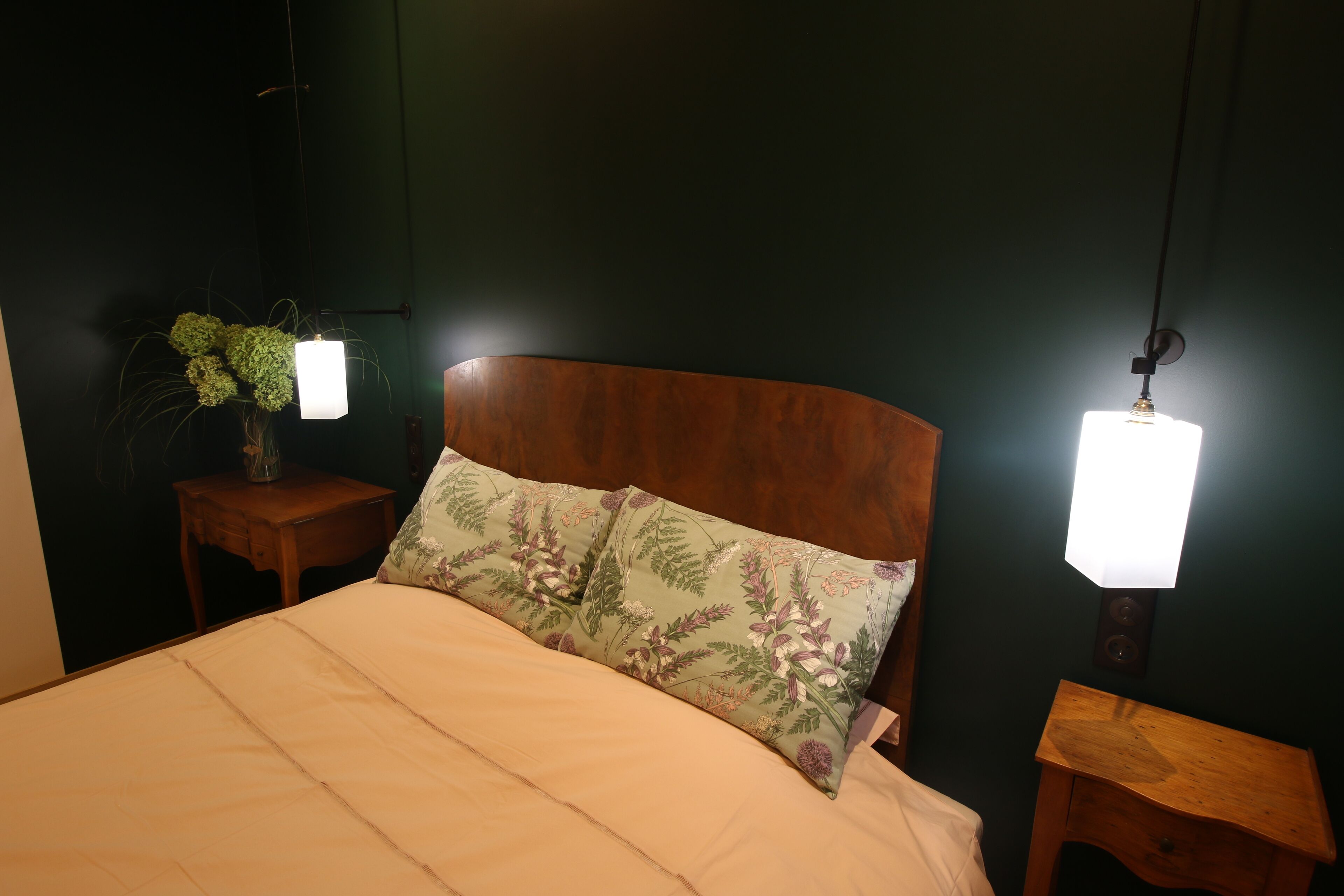 A bedroom with a wooden headboard, a bed with a beige bedspread, two bedside tables with lamps, and a wall-mounted plant.