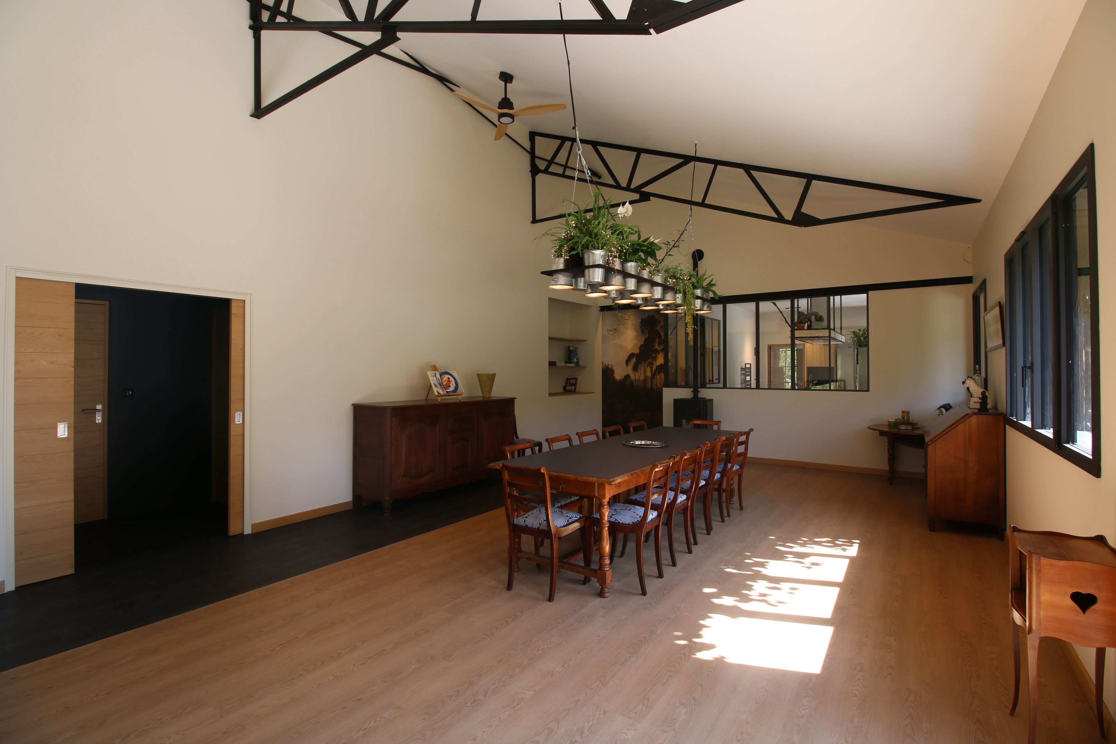 Un vaste espace dédié à la salle à manger, avec une longue table en bois et des chaises, un ventilateur de plafond et des plantes suspendues.