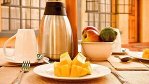 Un desayuno con una cafetera de acero inoxidable, una taza blanca, un bol de fruta y un plato de mango en rodajas.