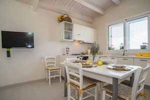A dining area with a white table set for four, white chairs, a television mounted on the wall, and a window with blinds. - VIlla White (Avola)