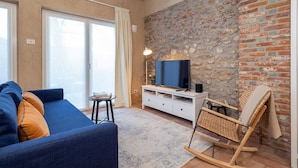 Een woonkamer met een blauwe bank, een televisie op een wit kastje, een stenen muur en een rieten schommelstoel.
