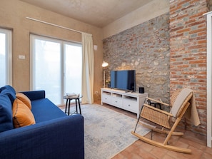 A living room with a blue sofa, a television on a white cabinet, a stone wall, and a wicker rocking chair.