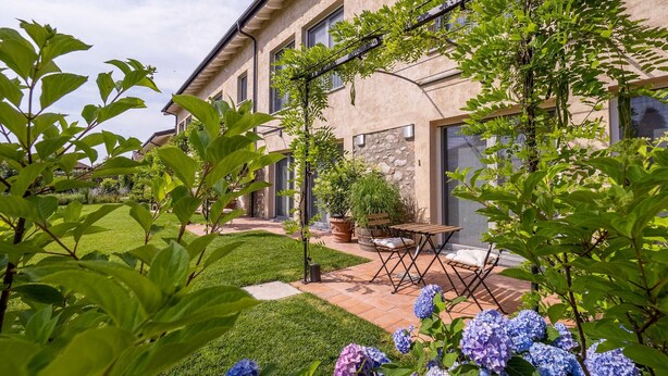 A well-maintained garden with hydrangea bushes, a patio with outdoor furniture, and a building with stone walls and large windows. - Peaceful Apartment with Garden in Farmhouse Resort (Cressa)