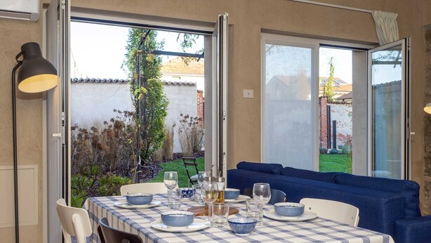 Een eetgedeelte met een tafel voor vier personen, een blauwe bank en uitzicht op een tuin via openslaande deuren.