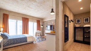 A bedroom with a bed, a desk, and a chair. There is a kitchen area with a counter and a television. The room has a stone wall and a window with curtains.