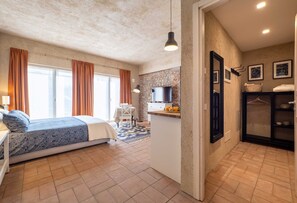A bedroom with a bed, a desk, and a chair. There is a kitchen area with a counter and a television. The room has a stone wall and a window with curtains.