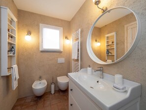 A bathroom with a white sink, bidet, and toilet, a round mirror, and a shelf with toiletries.