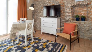 A cozy room with a stone wall, a flat-screen TV, a dining table with chairs, a white dresser, and two potted plants. - Quiet Studio Apartment with Garden in Farmhouse (Cressa)