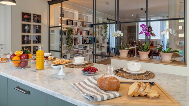 A modern kitchen with a marble countertop, a variety of fruits, pastries, and bread, and a view of a dining area through large glass doors.