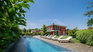 A swimming pool with wooden deck and lounge chairs, surrounded by greenery and a red building in the background.