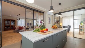 A modern kitchen with a marble countertop, a central island with fresh produce, and a dining area visible through glass doors.
