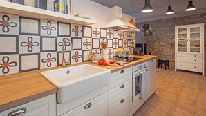 Una cucina moderna con mobili bianchi, un piano di lavoro in legno e una parete protettiva con motivi geometrici. Si notano lampade a sospensione, un forno integrato e una parete in pietra sullo sfondo.