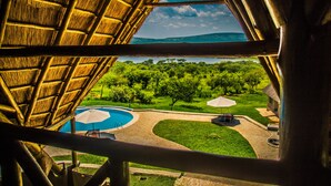 Vue depuis une chambre dont le toit est recouvert de chaume, donnant sur un espace piscine agrémenté de parasols et un jardin aménagé.