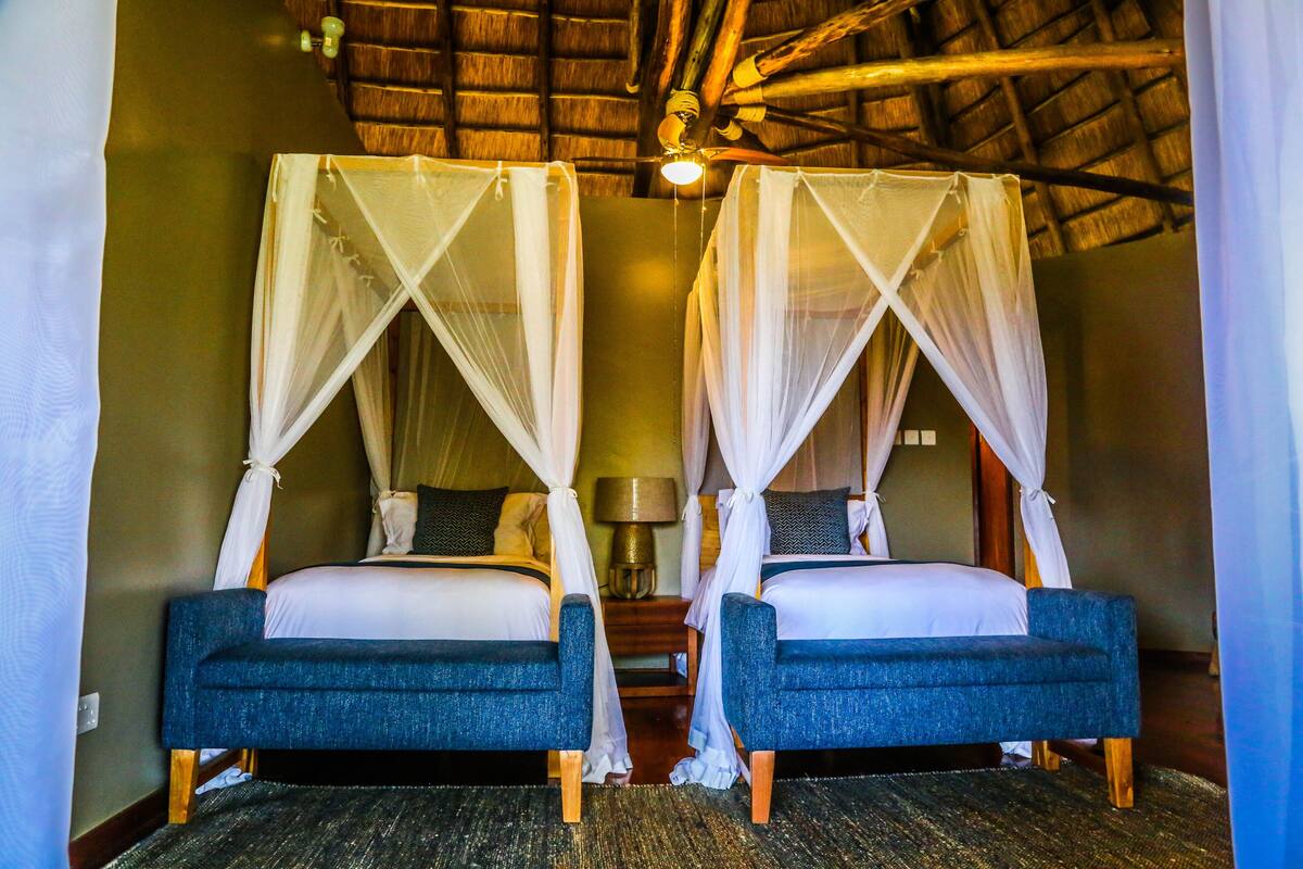 Two single beds with canopy beds, a wooden ceiling, and a nightstand with a lamp between them.