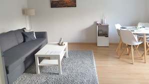 A living room with a grey sofa, a white coffee table, a small white side table with a lamp, a dining area with a white table and chairs, and a framed picture on the wall. - Pure relaxation with Nina (Bad Lauterberg im Harz)