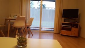 A living room with a dining table, two chairs, a glass vase with a plant, a television on a wooden stand, and a sliding glass door leading to a balcony. - Pure relaxation with Nina (Bad Lauterberg im Harz)