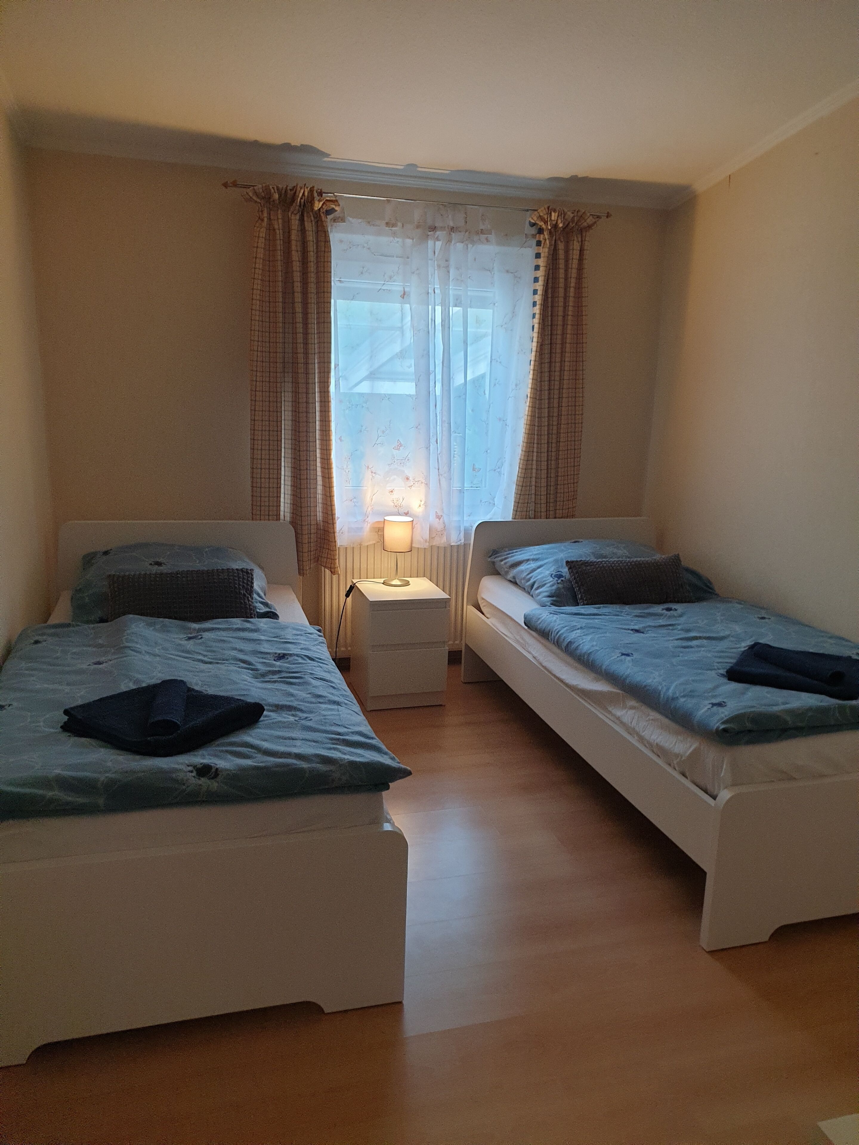 Une petite chambre bien éclairée, avec deux lits simples, une table de chevet avec une lampe et une fenêtre ornée de rideaux légers.