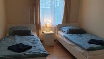 Une petite chambre bien éclairée, avec deux lits simples, une table de chevet avec une lampe et une fenêtre ornée de rideaux légers.