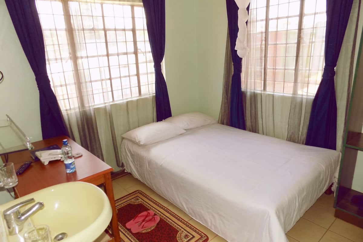 A bedroom with a bed, a bedside table with a lamp, a window with curtains, and a bathroom with a sink and mirror.