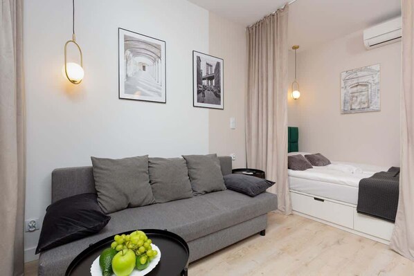 A modern living room with a grey sofa, a small black side table with a fruit bowl, a bed with a green headboard, and two framed pictures on the wall.