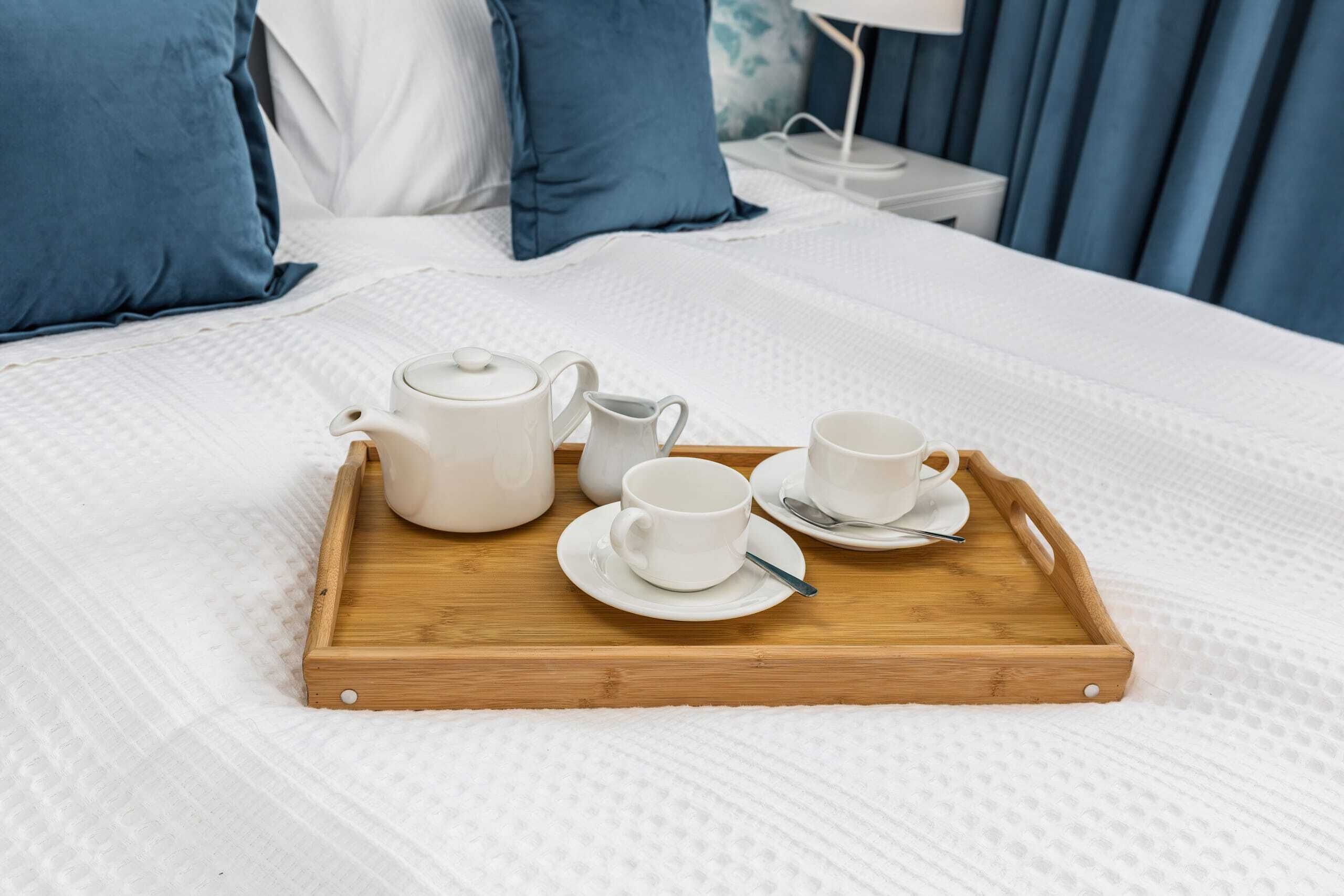 Ein ordentlich bezogenes Bett mit einem hölzernen Tablett, auf dem weiße Tassen und ein Teekessel stehen, auf einer weißen, strukturierten Bettdecke.