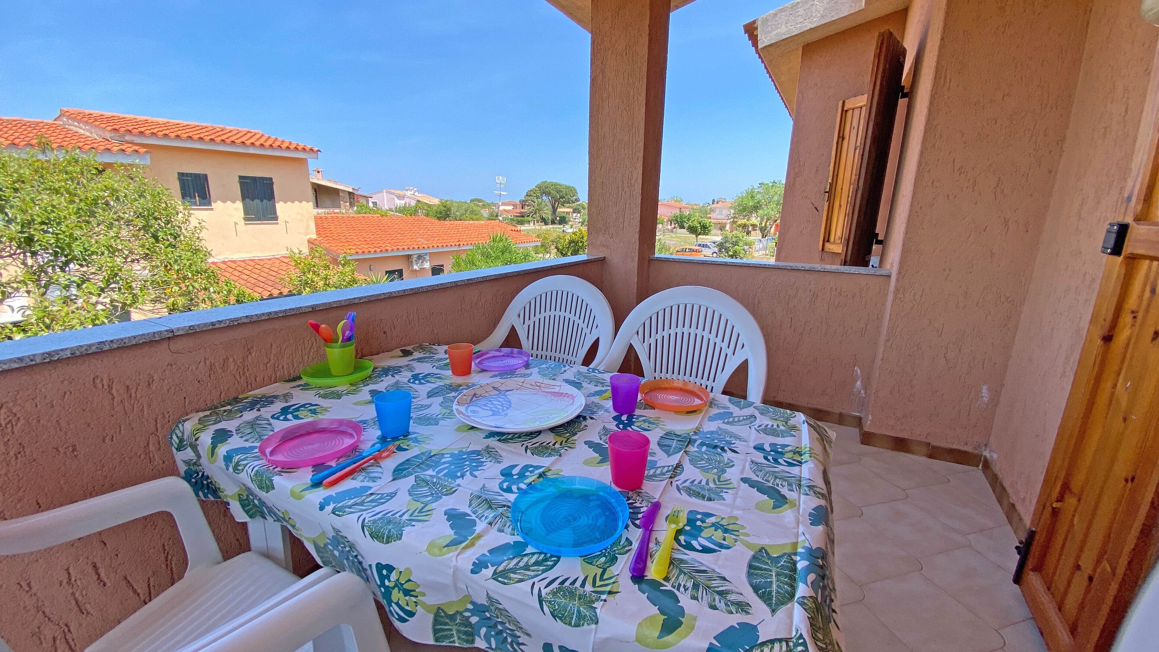 A balcony with a table set for dining, white chairs, and a view of buildings and trees.