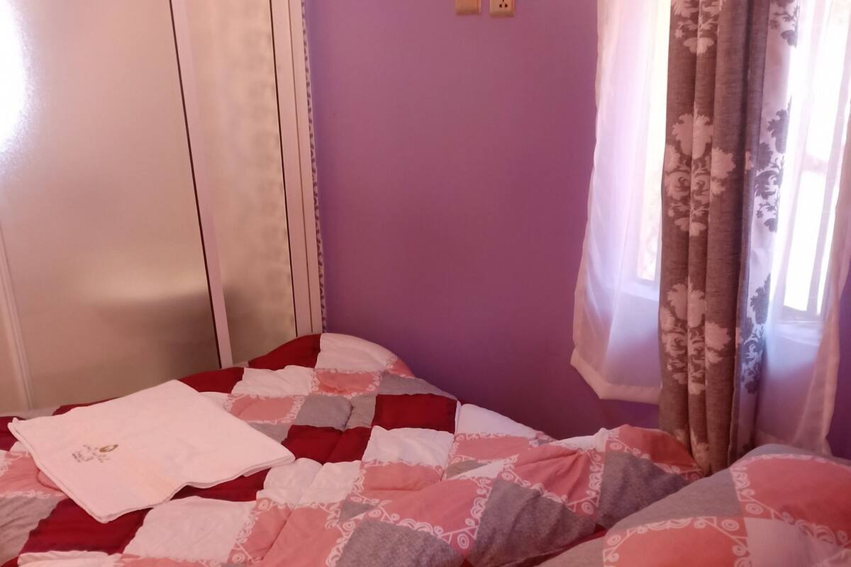 Una habitación con cama, cabecera de madera, ventana con cortinas y un enchufe en la pared.