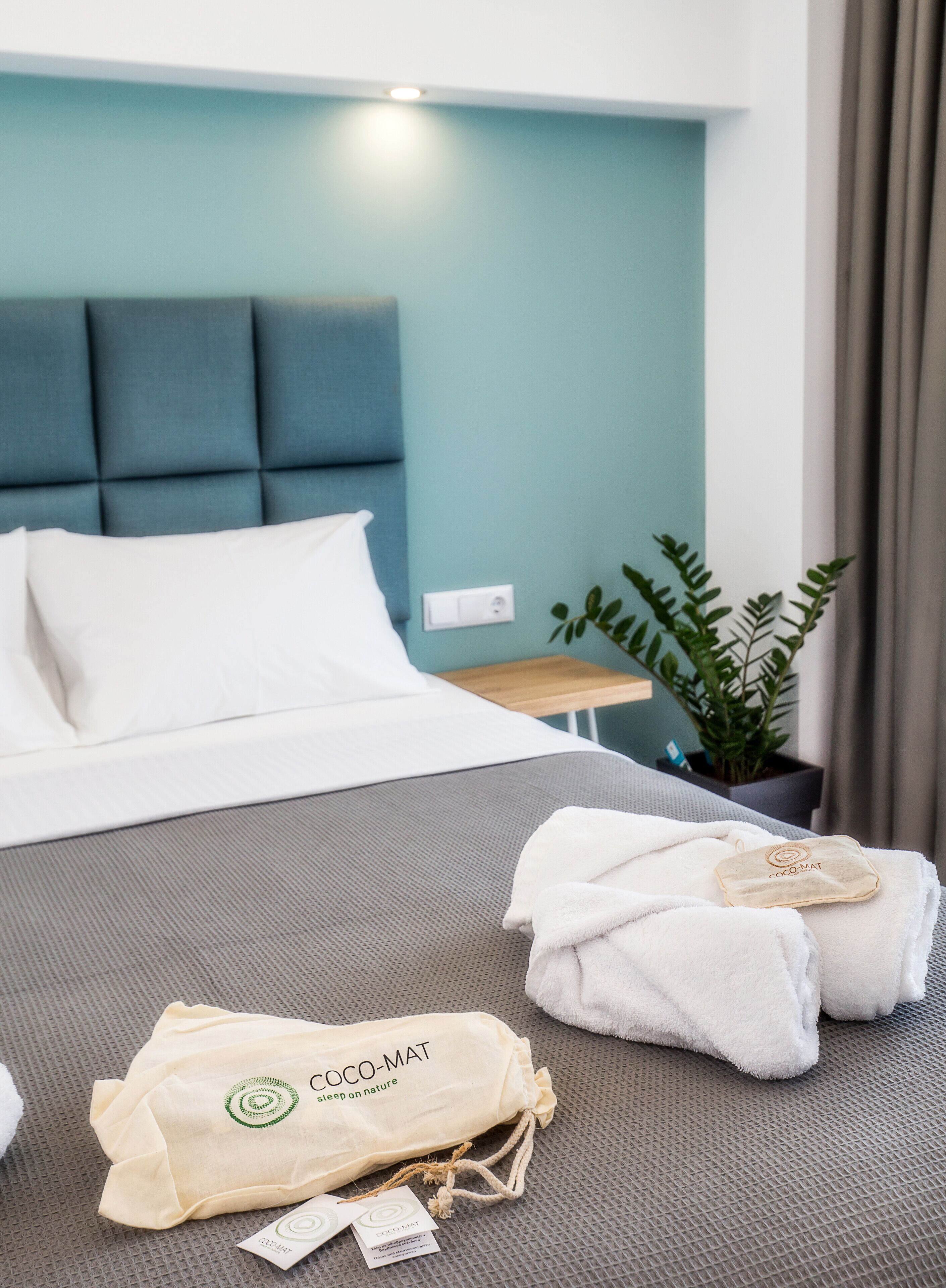 Ein Hotelzimmer mit einem Bett, einem kleinen Tisch, einer Topfpflanze sowie Handtüchern und einer Tasche mit dem Aufdruck "COCO-MAT".