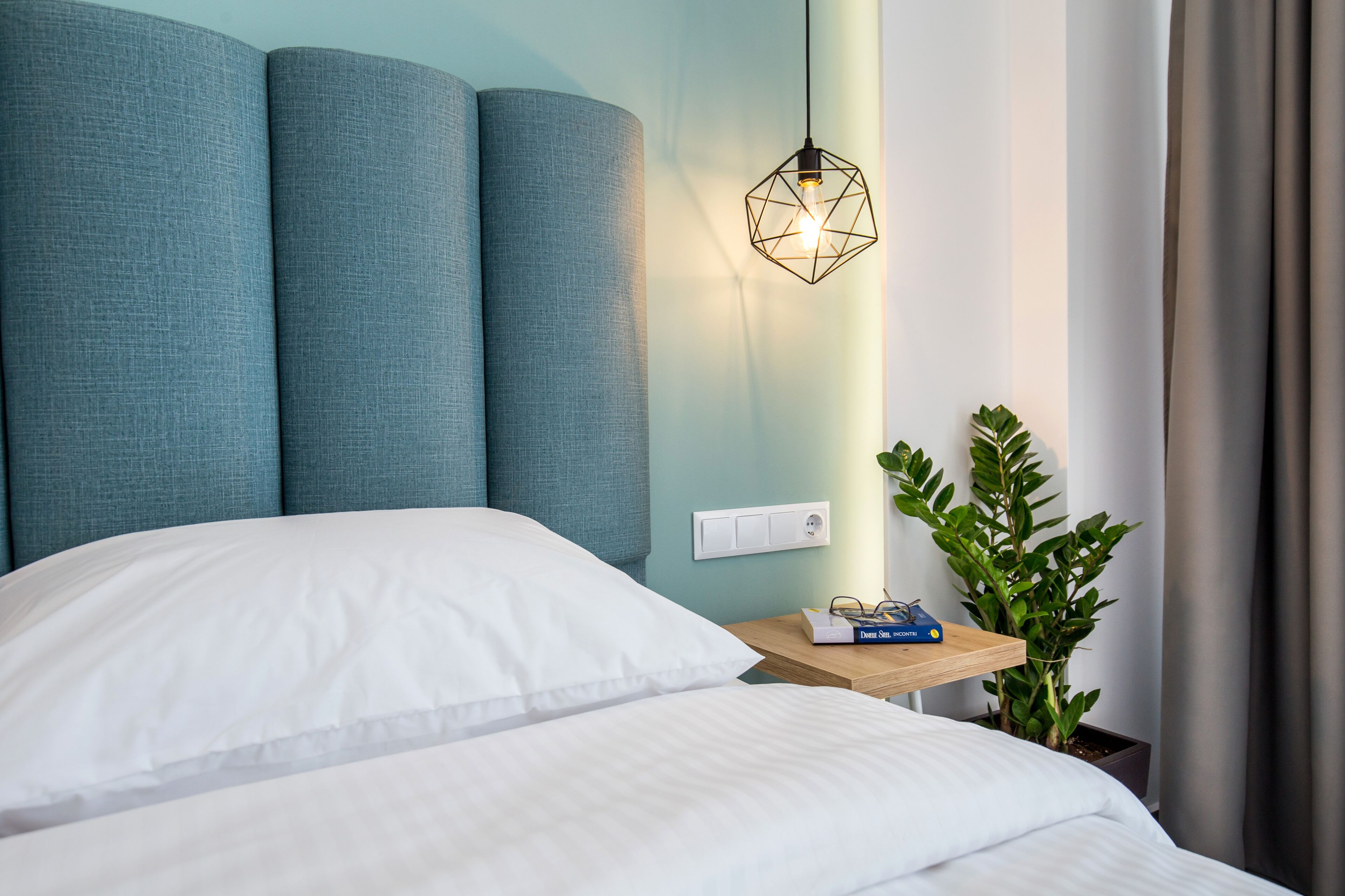 A neatly made bed with white linens, a geometric pendant light, a small bedside table with a book, and a potted plant.