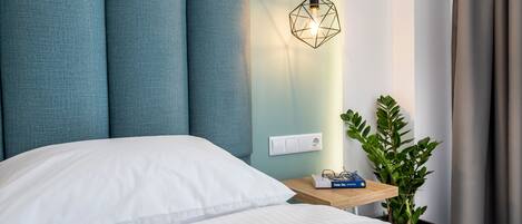 A neatly made bed with white linens, a geometric pendant light, a small bedside table with a book, and a potted plant.