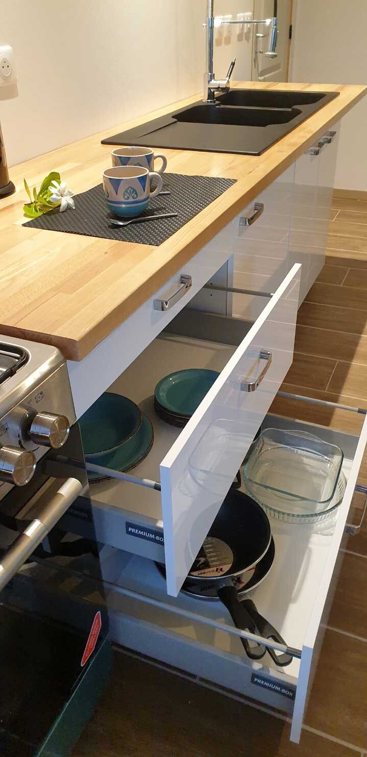 Eine Küche mit einer Holzausziehtafel, einem Spülbecken und einem Herd. Unter der Arbeitsplatte befinden sich zwei Schubladen, in einer davon liegen eine Glasschüssel und eine Bratpfanne.