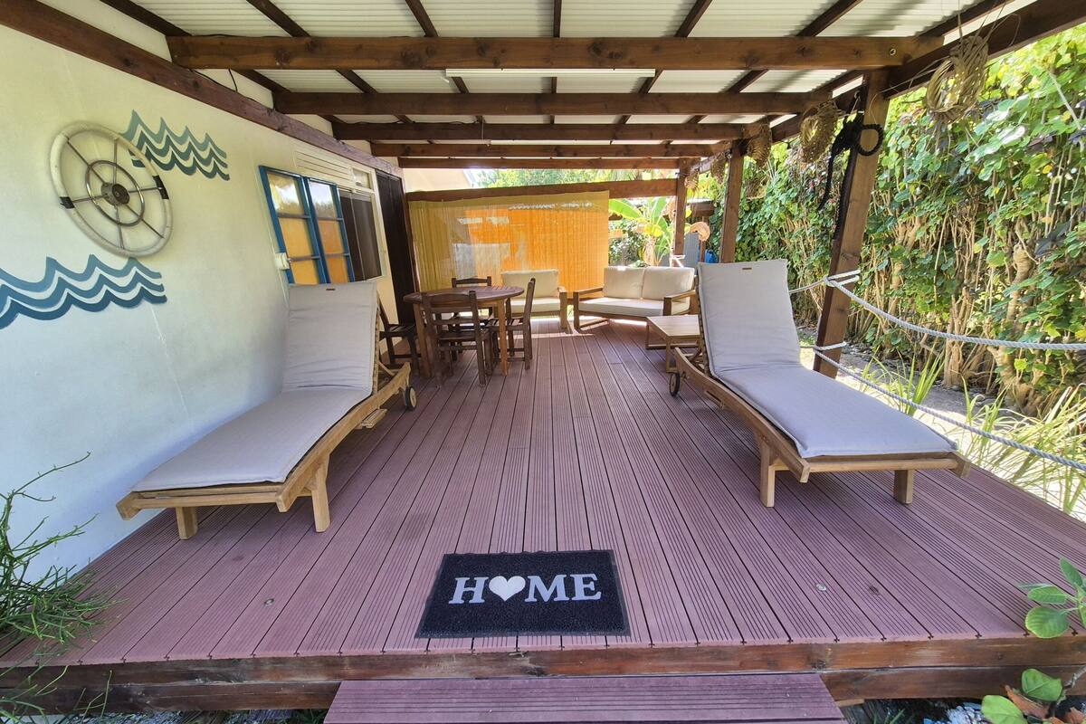 A wooden deck with lounge chairs and a table, a 'HOME' mat, and a wall with nautical decorations.