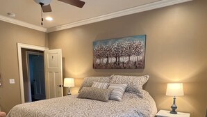 A bedroom with a bed, two bedside tables with lamps, a ceiling fan, a door, and a painting on the wall.