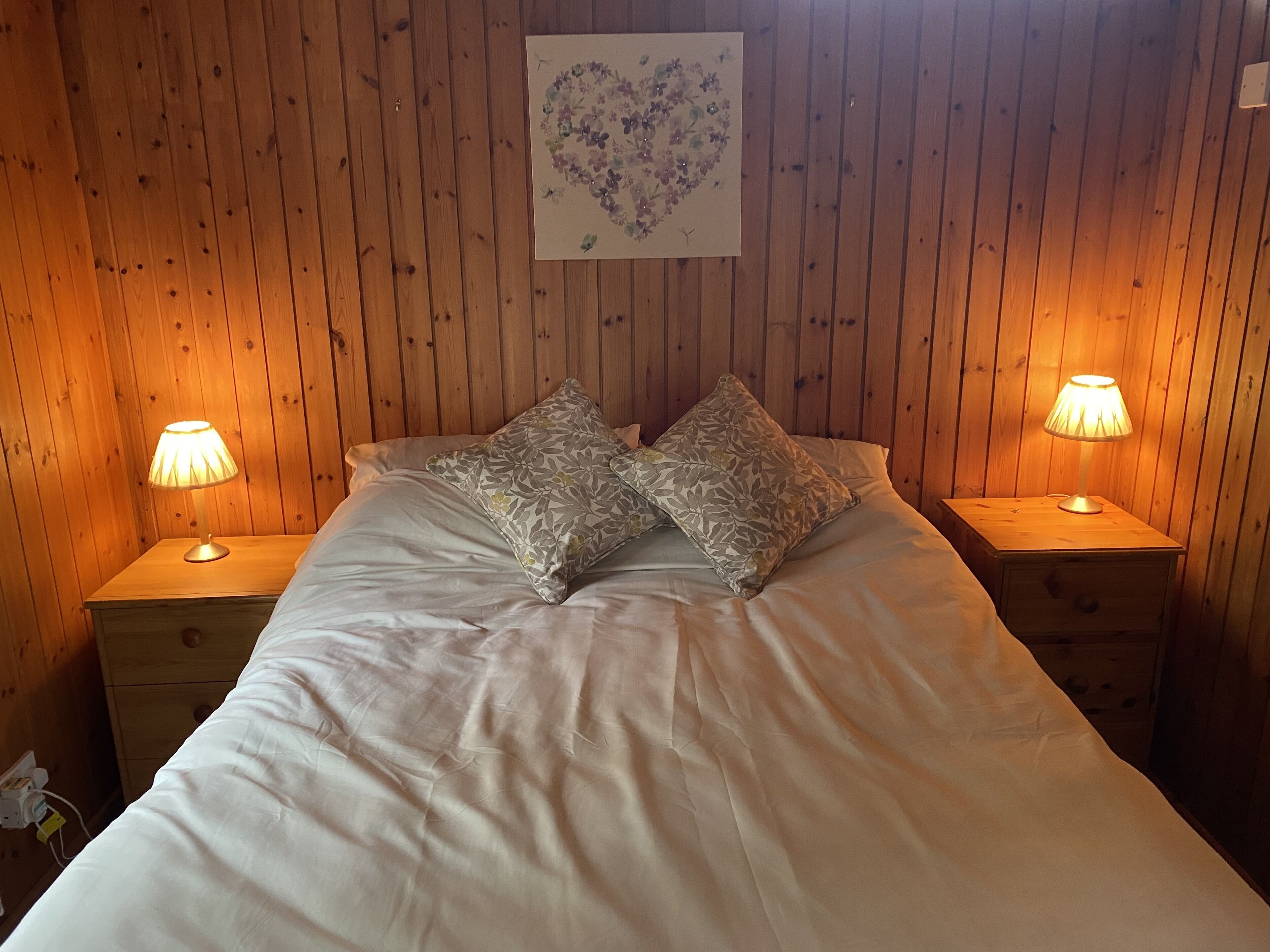 A wooden-paneled room with a bed, two bedside lamps, and a heart-shaped artwork on the wall.