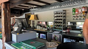 En bar med træloft, kasseapparat, computerskærm og hylder med flasker og glas.