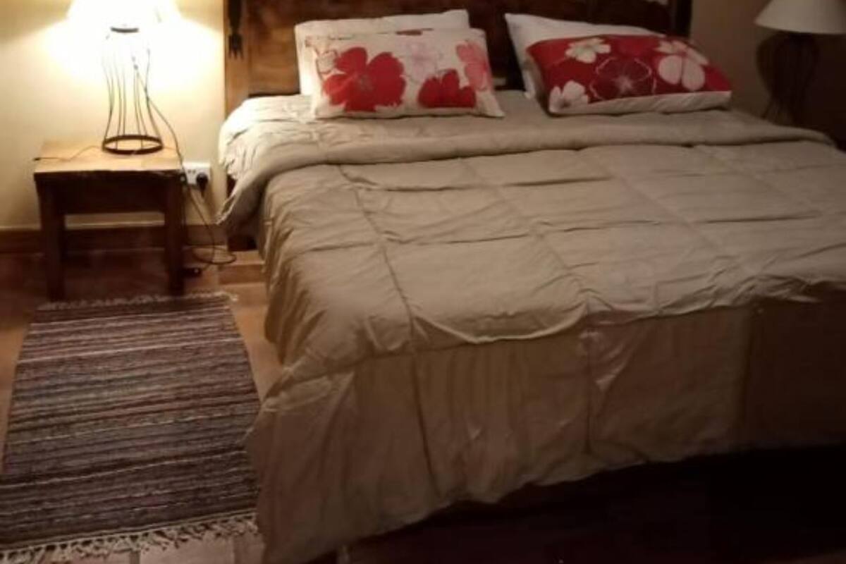 A bedroom with a wooden headboard, a bed with a beige duvet, two red floral pillows, a bedside table with a lamp, and a patterned rug.