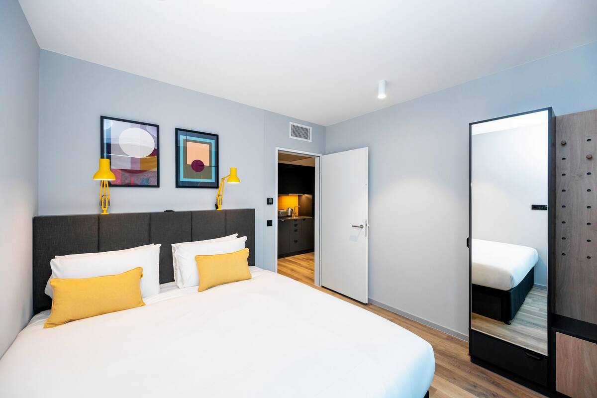 A modern hotel room with a large bed, a wardrobe, and a small kitchenette.